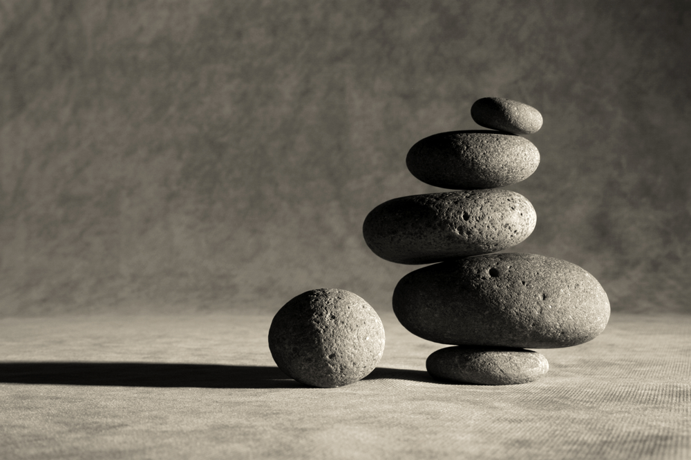 Smooth flat stones stacked as a carne on a soft grey background. There is a small spherical stone sitting by the shadow they cast.