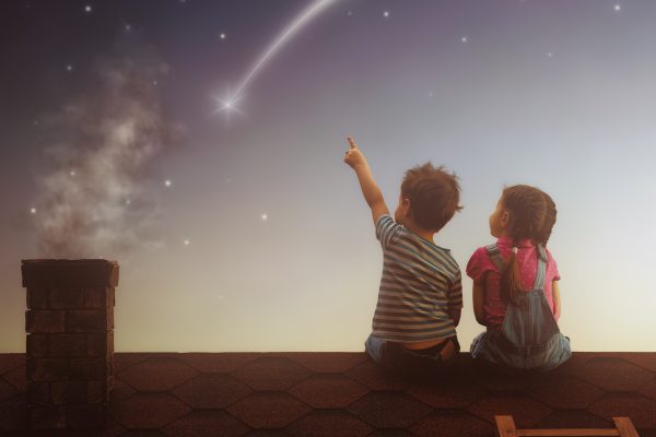 Two cute children sit on the roof and look at the stars. Boy and girl make a wish by seeing a shooting star.